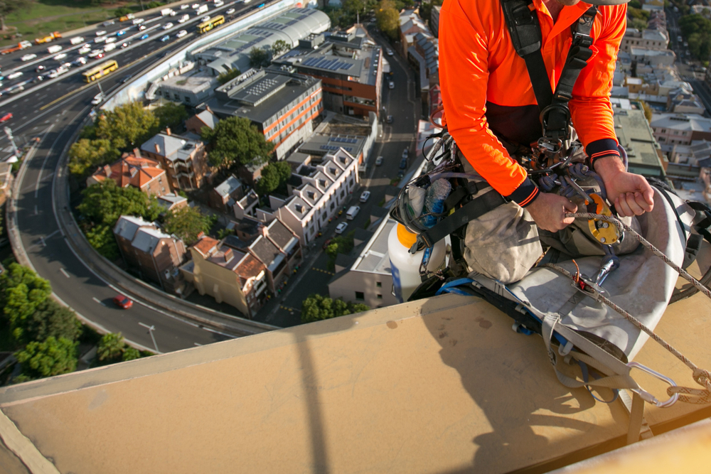 Worker caring for commercial roof using abseiling equipment.